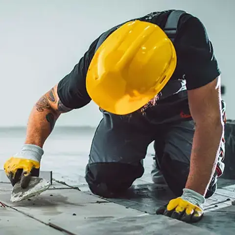 Contractor installing floor tile using a trowel while kneeling on a flooring surface and wearing protective safety gear.